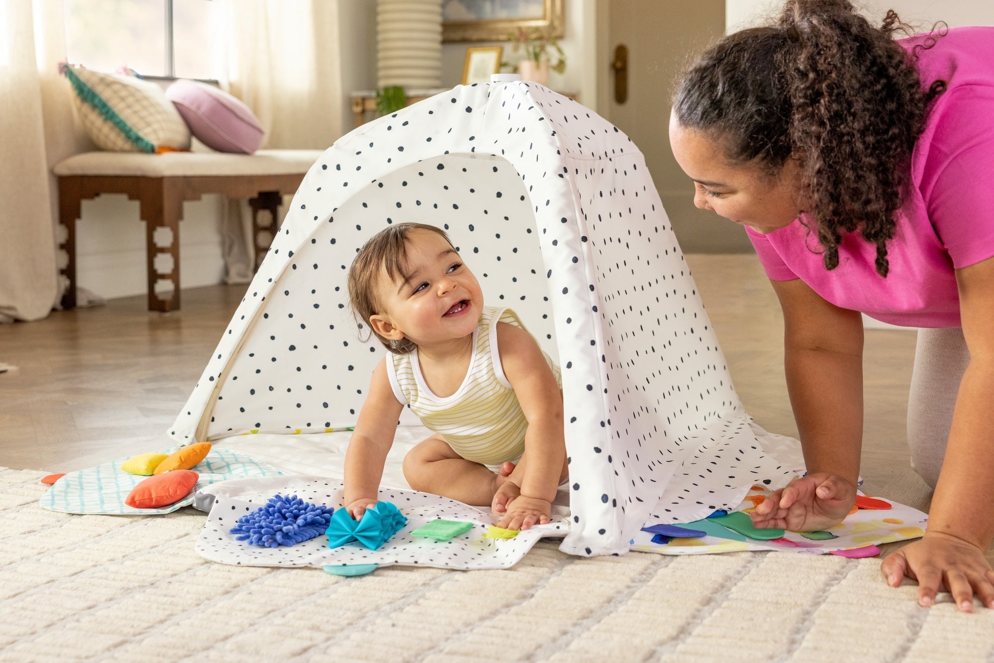 Baby playing on a mat with colorful toys under a white canopy, watched by an adult.