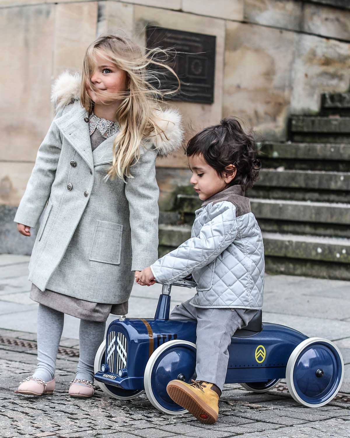 Two children playing with a blue bagera speedster ride-on toy car on a stone pavement.