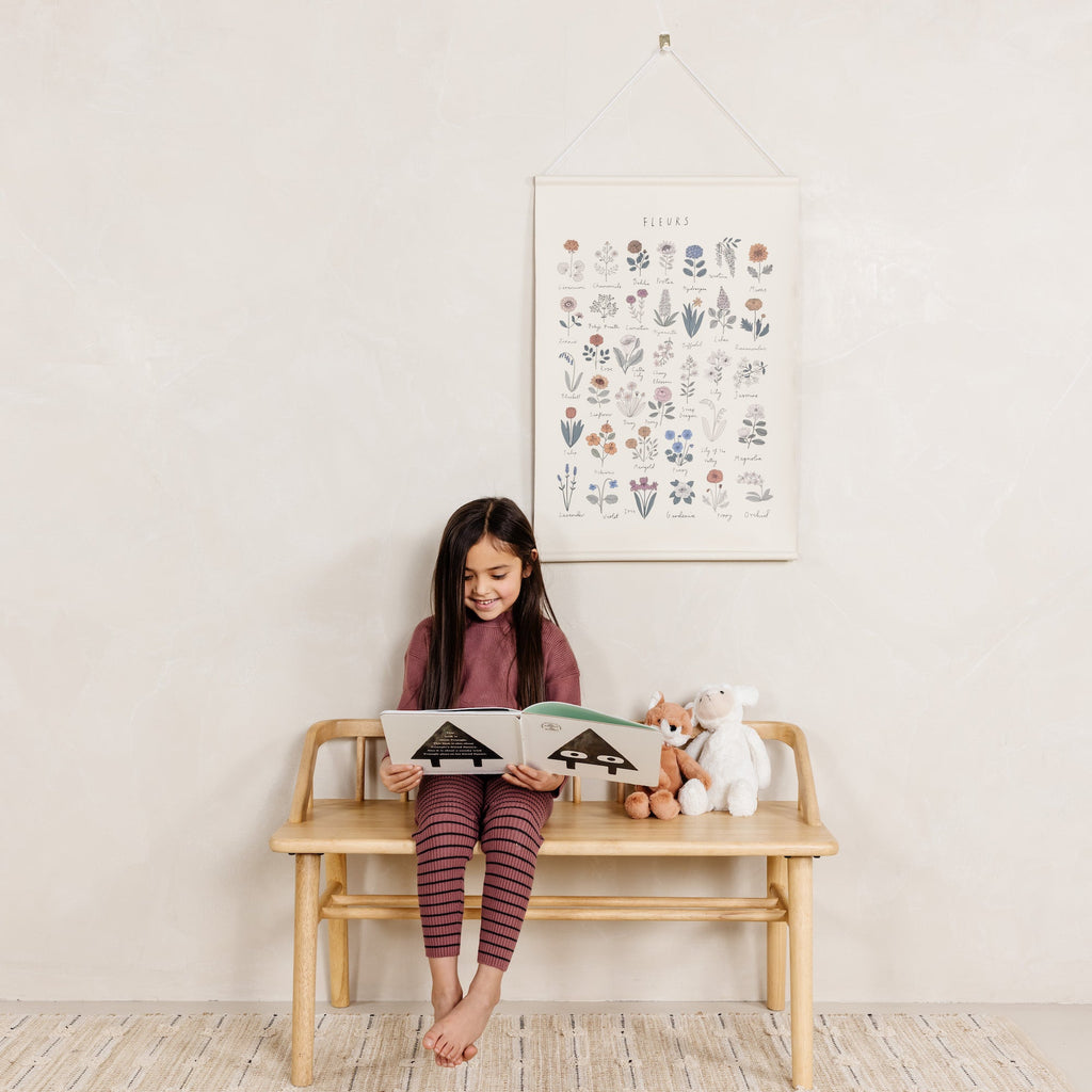 Young girl sitting on a wooden bench reading a book with a stuffed animal, in a room with a floral poster on the wall.