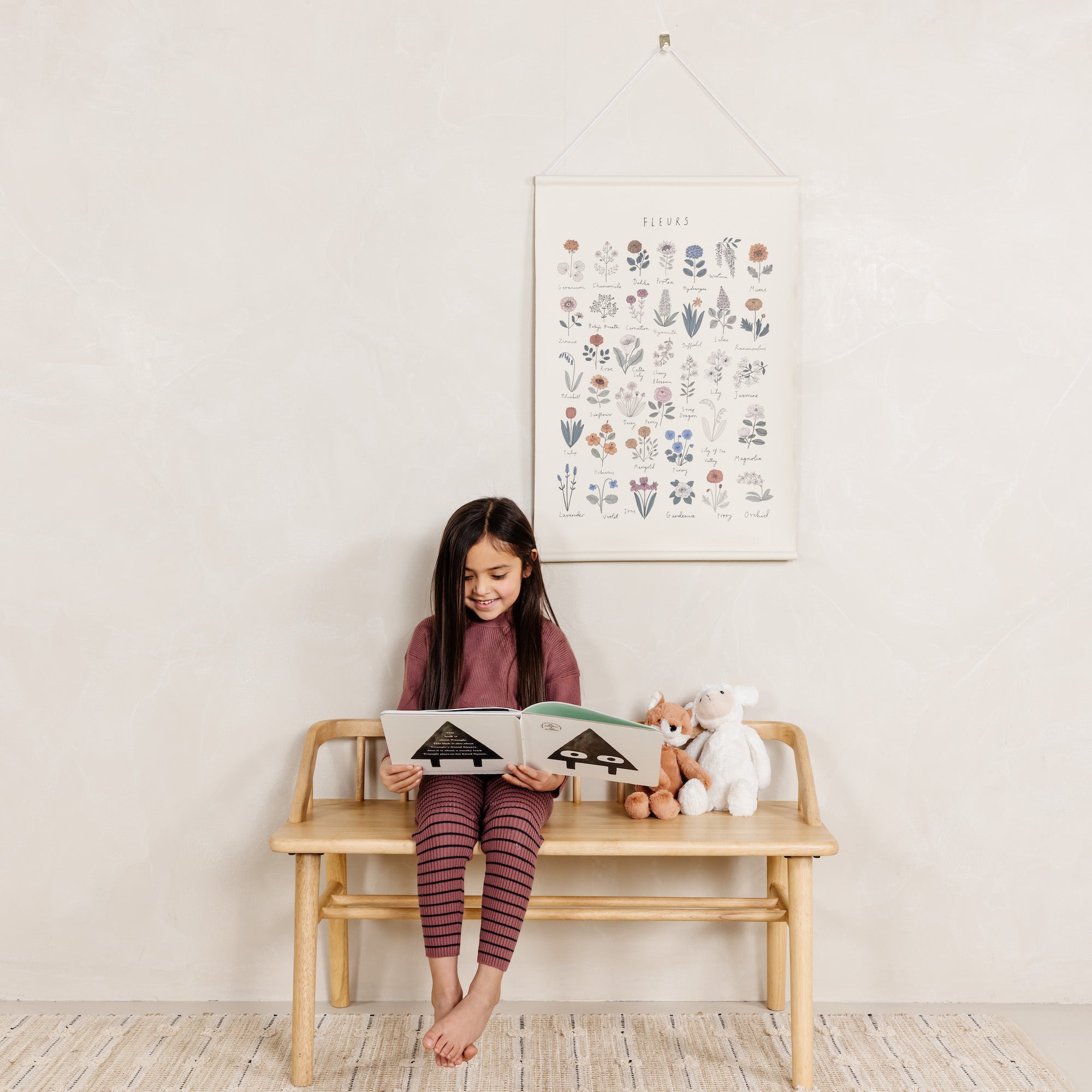 Young girl sitting on a wooden bench reading a book with a stuffed animal, in a room with a floral poster on the wall.