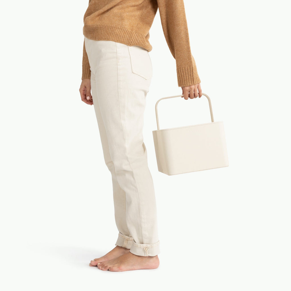 Person wearing a brown sweater and white pants holding a beige gathre cady on a white background