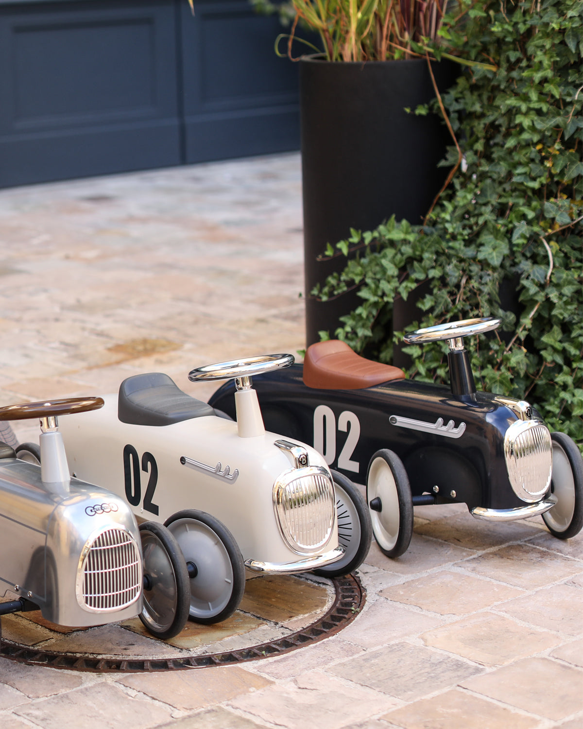 Two vintage-style baghera children's ride-on cars on a stone patio with plants in the background.