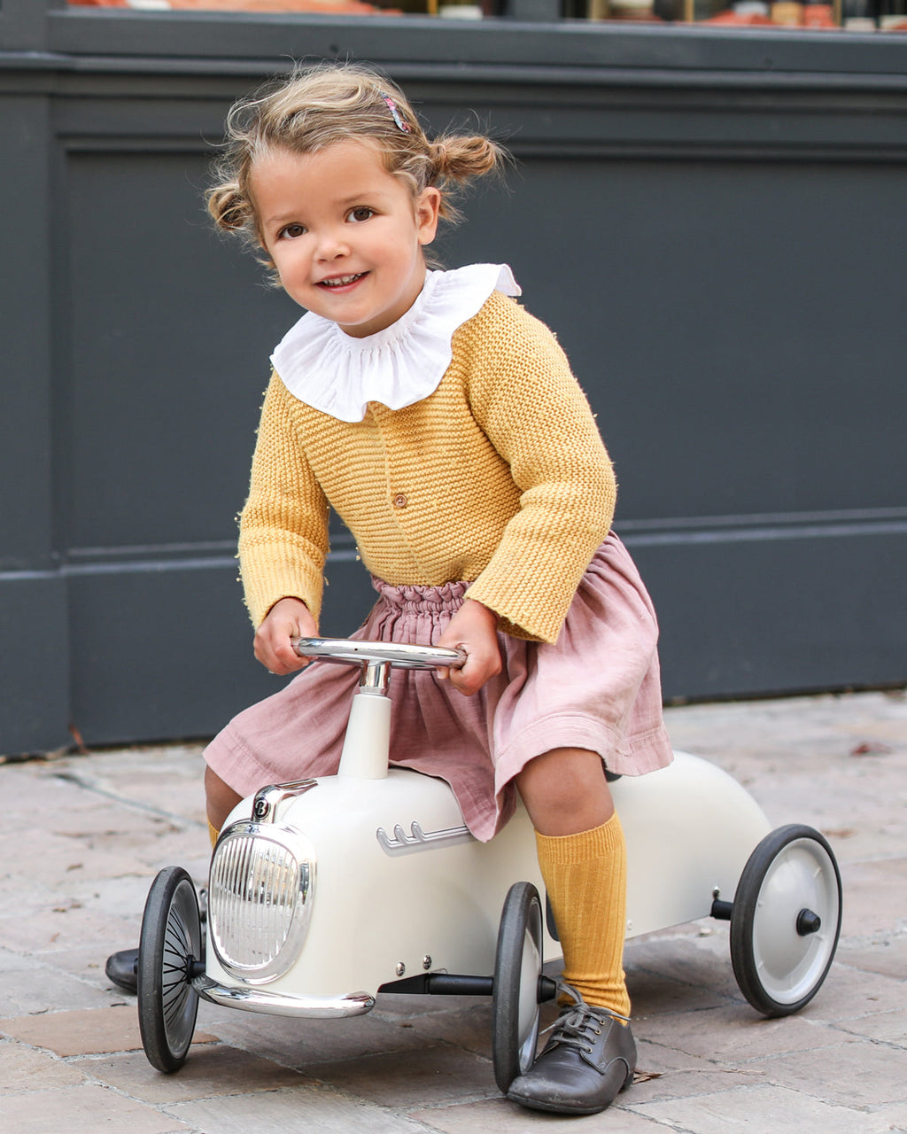 Child in a yellow sweater and pink skirt riding a toy car outdoors.
