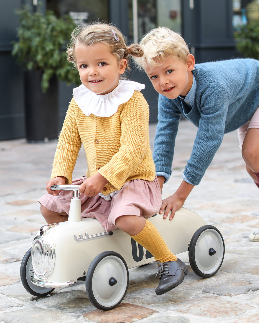 Two children playing with a toy car on a paved surface.