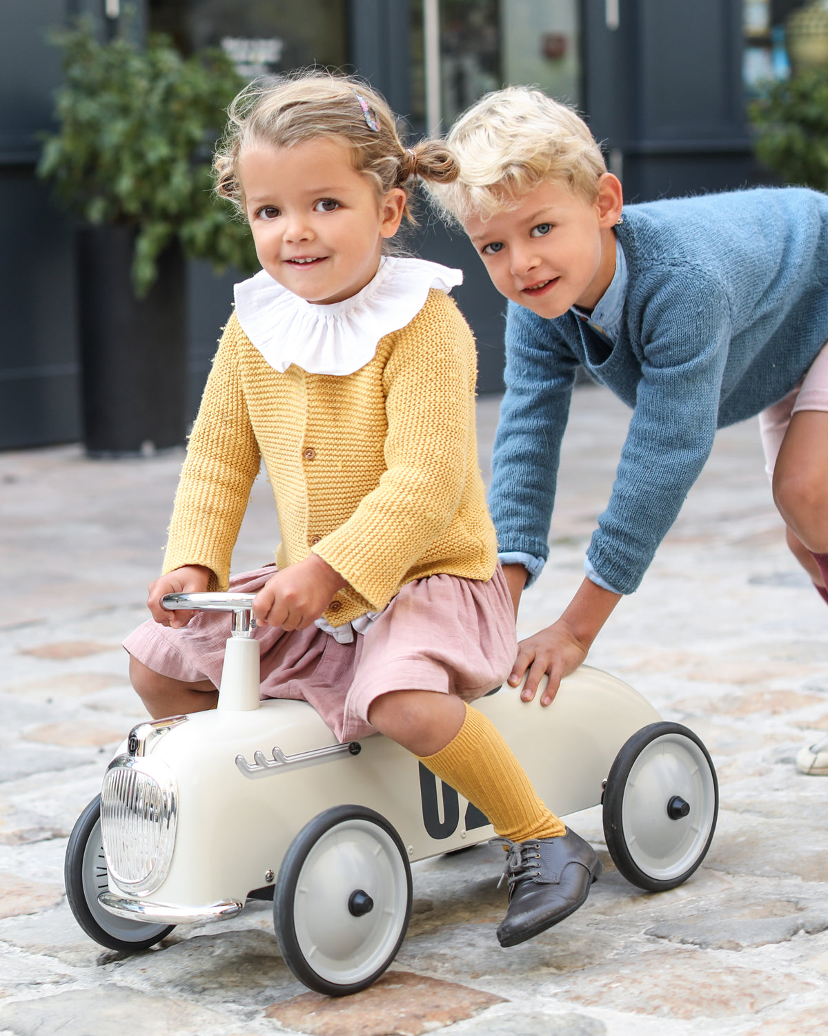 Two children playing with a toy car on a paved surface.