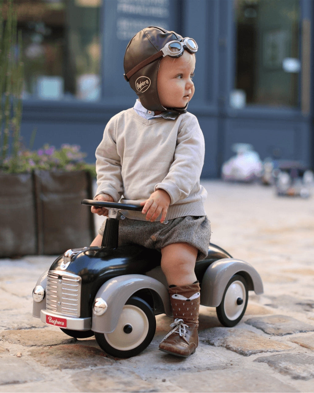Child riding a toy car wearing a helmet with goggles outdoors.