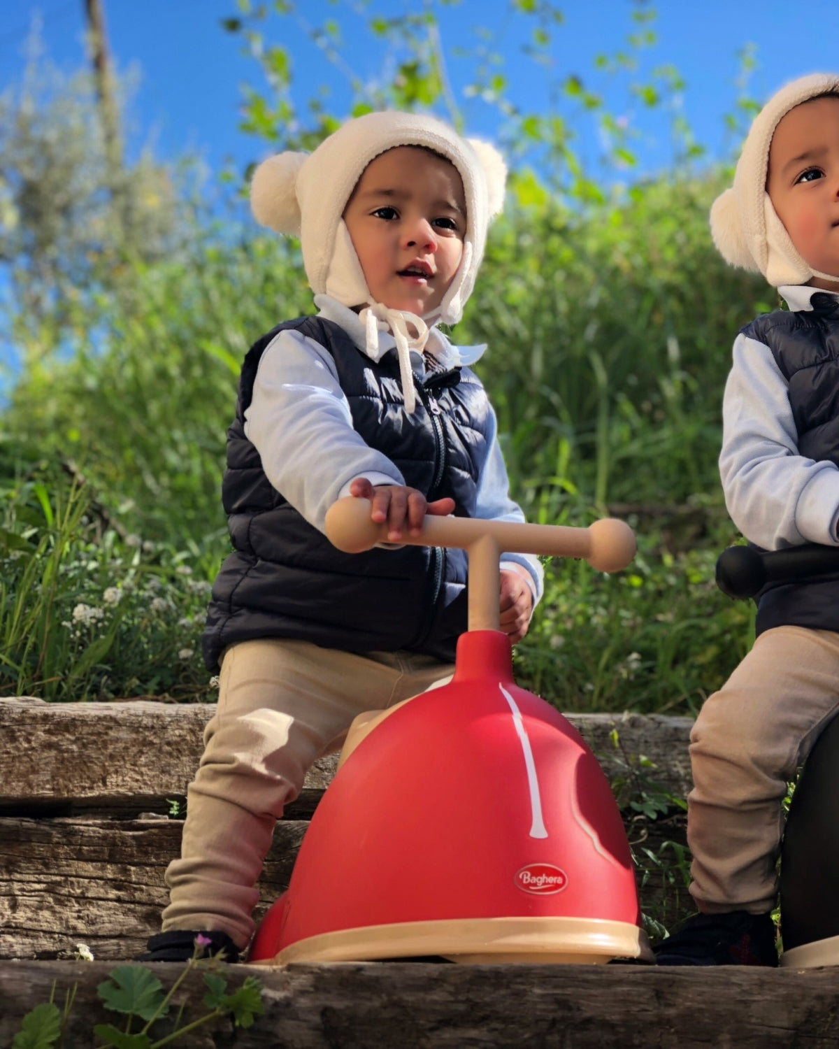Two children in winter clothing on a red and beige baghera speedster car outdoors.