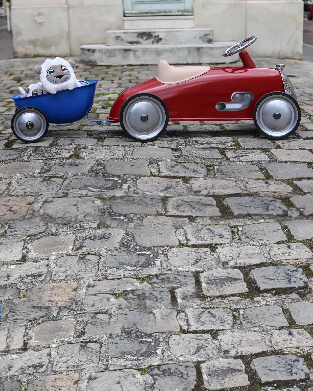 Red toy car with a blue trailer carrying a white dog on a cobblestone street.