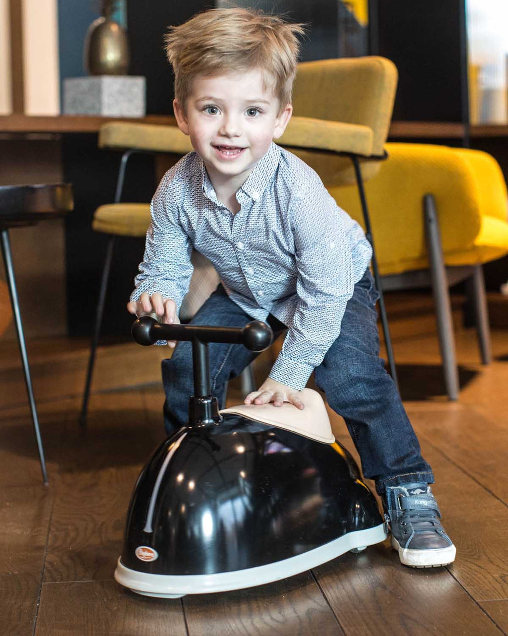 Child playing on a black ride-on  twister from baghera in a room with yellow chairs.